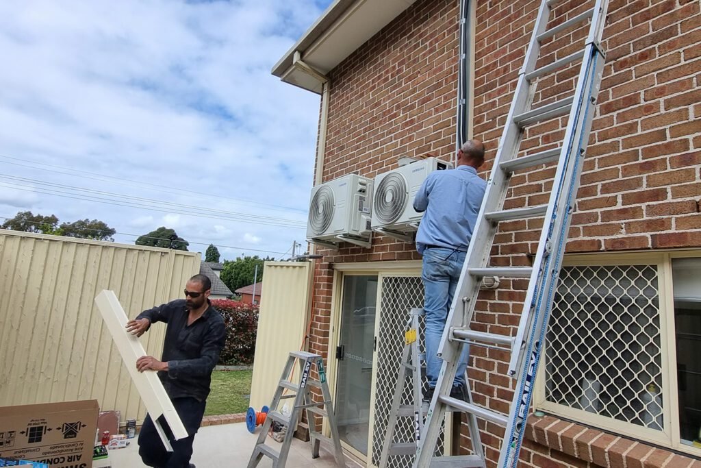 Technicians installing a reverse cycle air conditioning system on the exterior wall of a house.
